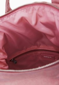 Interior de bolsa tote de tela rosa con forro liso y brillante y un bolsillo con cremallera. La etiqueta del bolsillo dice "Desigual." Cierre superior con cremallera.