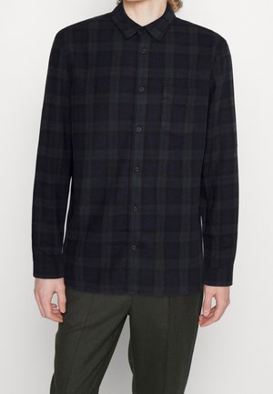Homme portant une chemise à carreaux sombre à manches longues et un pantalon foncé, debout devant un fond clair uni.