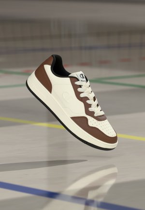 Bílá a hnědá teniska s perforovanou špičkou, hladkým koženým svrškem, krémovou stélkou, černou pryžovou podrážkou a logem na jazyku.