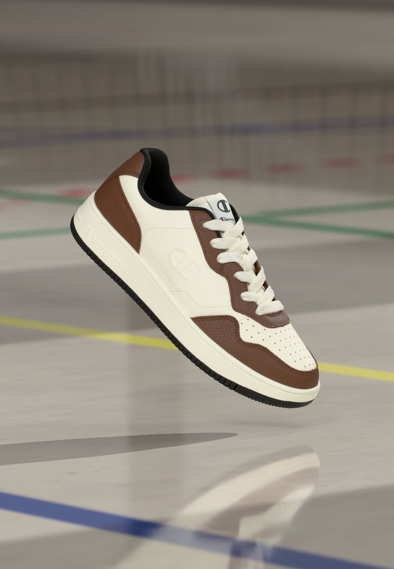 Λευκό και καφέ αθλητικό παπούτσι με διάτρητο μύτη, λείο δερμάτινο επάνω μέρος, μπεζ μεσαία σόλα, μαύρη λαστιχένια σόλα και λογότυπο στη γλώσσα.