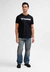 Ung man stående, iförd en svart t-shirt med "hummel"-logotyp, blå jeans och svarta sneakers mot en enkel vit bakgrund.