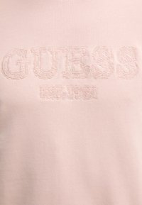 Sudadera de un suave color rosa pálido con bordado en relieve que presenta "GUESS" y "EST. 1981" en un color de hilo contrastante. Tejido suave y texturizado.