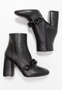 Botas de tobillo de cuero negro con un tacón grueso, que cuentan con un detalle de cadena en la parte superior y una cremallera lateral para el cierre. Textura suave.
