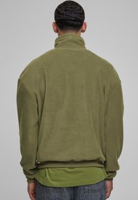 Pullover in pile verde oliva con colletto alto, vestibilità ampia, polsini e orlo a coste; texture morbida con dettagli di cucitura minimi.