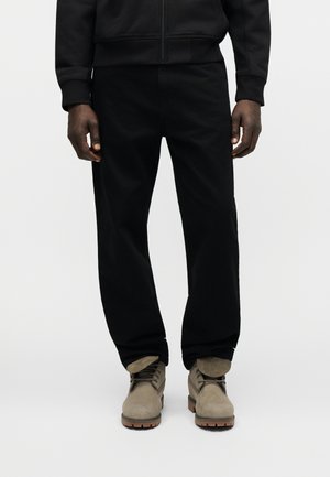 Muškarac u crnim hlačama i crnoj jakni stoji, nosi bež antilop čizme s vezicama i gumenim potplatima.