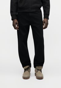 Άνδρας με μαύρο παντελόνι και μαύρο μπουφάν, που στέκεται, φοράει μπεζ δερμάτινα μποτάκια με κορδόνια και καουτσούκ σόλες.