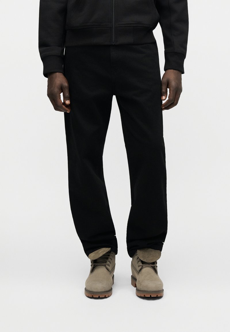 Mann in schwarzen Hosen und schwarzer Jacke stehend, trägt beige Wildleder-Schnürstiefel mit Gummisohlen.