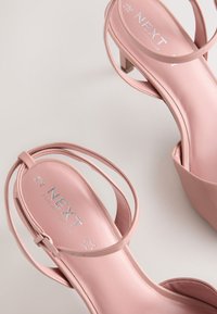 Sandales à talons hauts roses avec une finition brillante, fines brides autour de la cheville et une semelle intérieure rembourrée portant le logo "NEXT" en argent. Design à talon bloc.