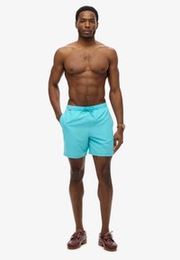 Shorts de bain turquoise avec une taille élastique et un cordon de serrage, arborant un petit logo sur le côté. Tissu lisse et léger.