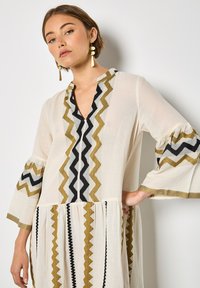 Tunique blanche avec des broderies en zigzag colorées en or, noir et gris. Présente des manches évasées et un décolleté en V, avec un ourlet à volants.