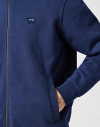 Felpa con zip di colore blu navy in morbido tessuto, con logo triangolare Wrangler, tasche laterali e polsini a costine. Design semplice, vestibilità casual.