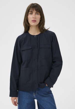 Woman wearing a navy button-up jacket with chest and lower pockets, paired with blue jeans, against a plain white background.