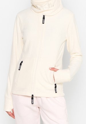 Woman wearing a cream-colored zip-up jacket with three visible black zipper pulls labeled "Bench," hand in right pocket.