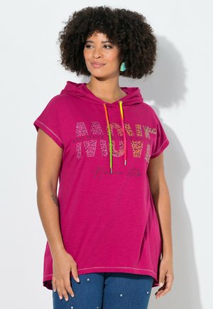 Kvinde med krøllet hår iført en magenta kortærmet hættetrøje med farverige snore og tekst, kombineret med blå jeans med små nitter.