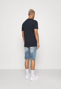 Mężczyzna z krótkimi blond włosami, ubrany w czarną koszulkę, wyblakłe niebieskie szorty dżinsowe, białe skarpetki i białe sneakersy, stojący tyłem.