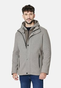 Hombre vistiendo un abrigo con capucha gris claro sobre una chaqueta marrón con cremallera y jeans azules, de pie contra un fondo blanco liso.