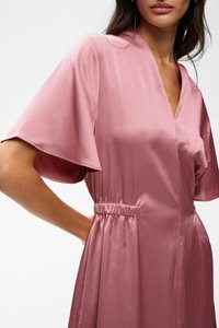 Femme portant une robe rose texturée avec de larges manches courtes et une taille élastique, montrée des épaules aux hanches sur un fond uni.