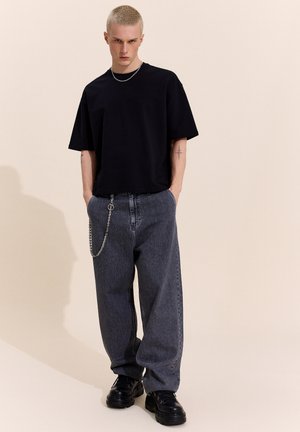 Mladý muž s krátkymi blond vlasmi, ktorý nosí čierne oversized tričko, voľné sivé rifle s reťazou a čierne masívne topánky, stojaci s rukami v vreckách.