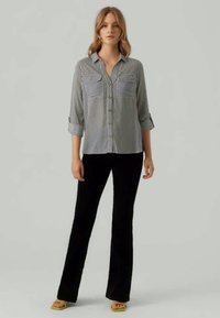 Gestreiftes Hemd mit zwei Brusttaschen und hochgerollten Ärmeln, kombiniert mit schwarzen Schlaghosen und gelben Sandalen. Glatte Stofftextur.