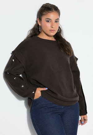 Ung kvinde med langt mørkt hår iført en brun sweater med metalhjerter på ærmerne og blå jeans, poserer med den ene hånd i lommen.