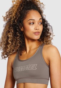 Jasnobrązowy stanik sportowy z okrągłym dekoltem, z wyraźnym białym logo "HUMMEL" na przodzie oraz gładką, elastyczną fakturą materiału.