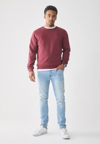 Young man standing wearing a maroon sweatshirt, light blue jeans, white sneakers, and a wristwatch against a plain white background.