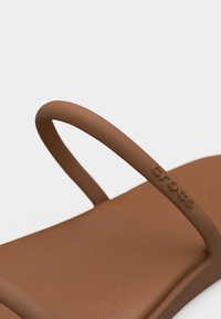 Καφέ σανδάλι Crocs με λεπτό λουράκι που φέρει το ανάγλυφο λογότυπο Crocs σε απλό φόντο.