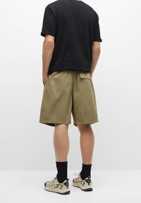 Shorts khaki avec une coupe décontractée, dotés de poches latérales et d'une taille à cordon de serrage, associés à un T-shirt noir et des chaussures de sport légères.