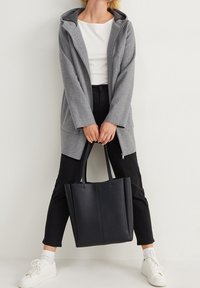 Personne portant un manteau à capuche gris, une chemise blanche, un pantalon noir et des baskets blanches, tenant un grand sac fourre-tout noir à deux mains.