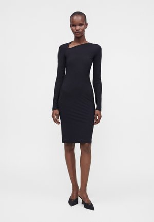 Femme portant une robe noire cintrée à manches longues longueur genou avec un décolleté asymétrique et des talons noirs pointus, debout sur un fond uni.