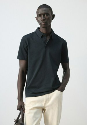 Hombre con camisa tipo polo de manga corta negra y pantalones beige, sosteniendo una bolsa negra en una mano, de pie contra un fondo liso y claro.