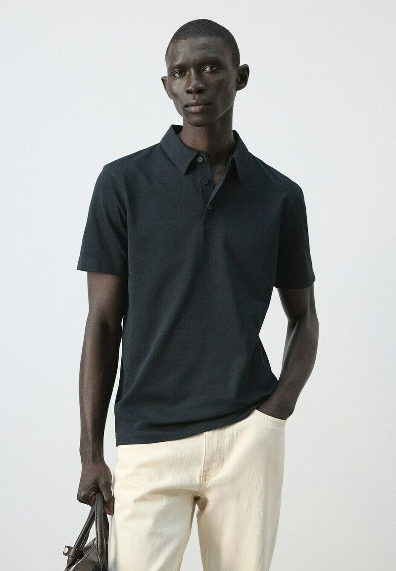 Homme portant un polo noir à manches courtes et un pantalon beige, tenant un sac noir dans une main, debout devant un fond clair uni.