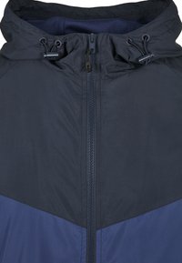 Marinfärgad vindjacka med dragkedja framtill, dragsko i huvan och kontrasterande mörkblå chevronmönster på den nedre delen. Slät, lättviktig tyg.