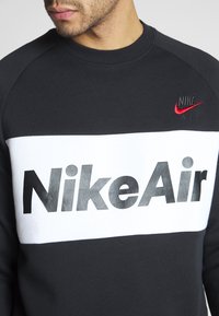Svart sweatshirt med vit bröstpanel, som har "Nike Air" i stora svarta bokstäver. Liten röd Nike-logga på vänster bröst. Tillverkad av bomull.