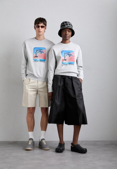 To modeller viser matchende grå sweatshirt med en delfin-illustrasjon i pastellfarger. Den ene har på seg beige shorts, mens den andre har svarte satengshorts, og de har forskjellige fottøy.