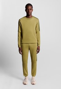 Olivgrünes Sweatshirt und Hosen Set, mit Rundhalsausschnitt, gerippten Bündchen und seitlichen Streifen. Getragen mit beigen Sportschuhen. Einfaches, tailliertes Design.