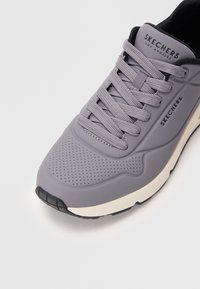 Harmaa Skechers-tenniskenkä nauhoilla, rei'itetty varvasalue, musta-valkoinen pohja, valkoista taustaa vasten.