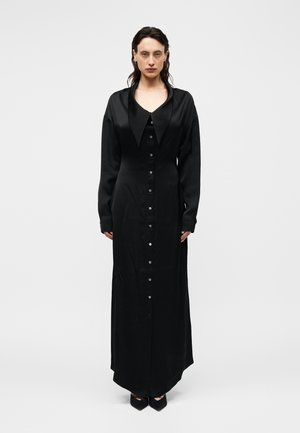 Femme debout, de face, portant une longue robe noire boutonnée avec un large col en satin et des manches longues sur un fond uni.