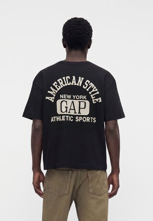 Muž otočený chrbtom, má oblečené čierne tričko s nápisom „American Style New York GAP Athletic Sports“ na chrbte a khaki nohavice.