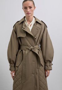 Brauner Trenchcoat mit Gürteldesign und gestepptem Muster sowie oversized Ärmeln. Verfügt über breite Revers, Druckknöpfe und seitliche Taschen.