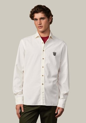 Camisa blanca de algodón con mangas largas, con botones en el frente y un pequeño logo negro en el pecho. Combinada con pantalones verde oscuro.