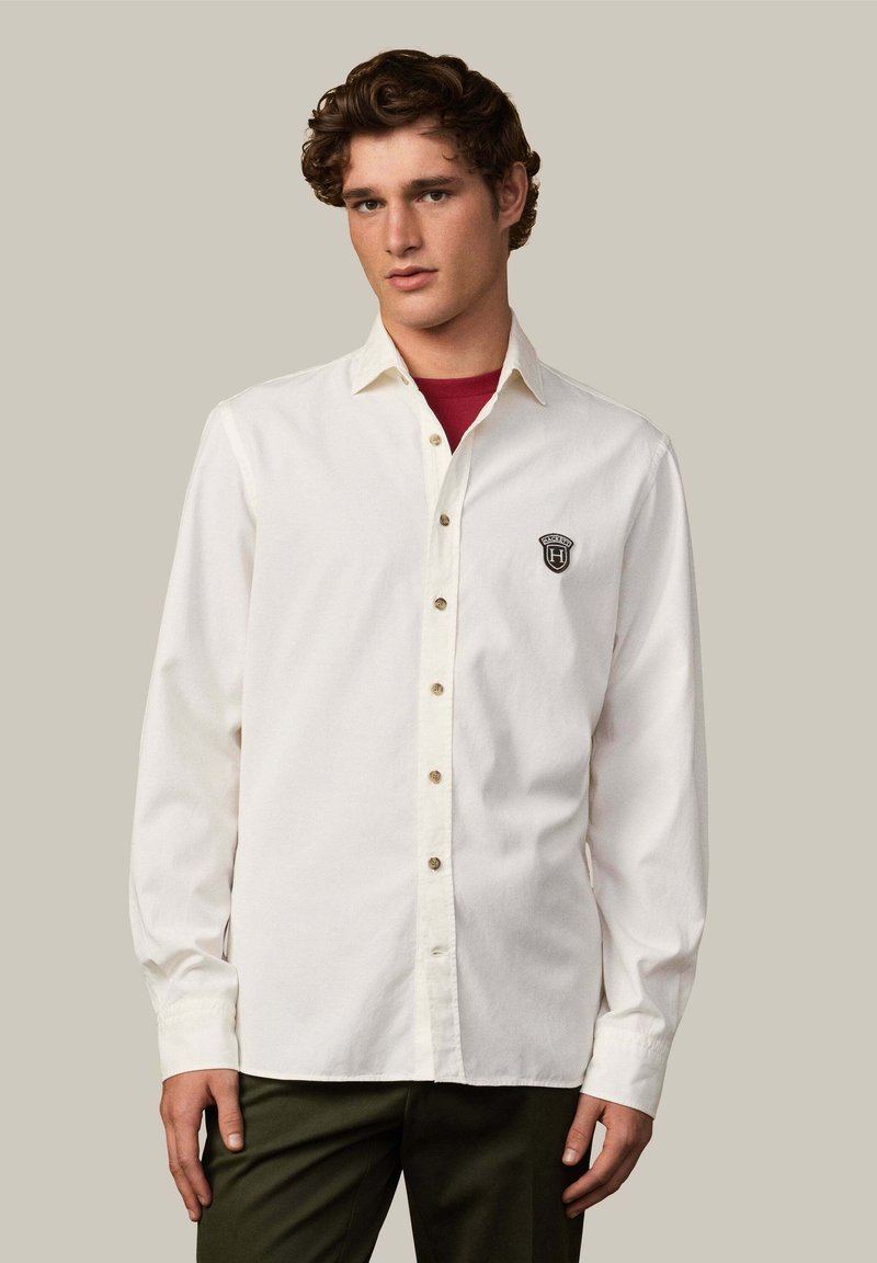 Camisa blanca de algodón con mangas largas, con botones en el frente y un pequeño logo negro en el pecho. Combinada con pantalones verde oscuro.