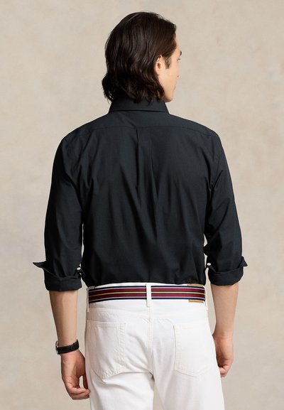 Moški s temnimi lasmi, oblečen v črno majico z dolgimi rokavi z zavihki, bele hlače in pas s črtami v rdeči, rumeni in mornarsko modri barvi, obrnjen stran.