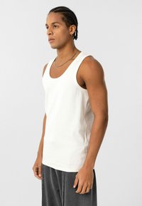 Man met gevlochten haar, gekleed in een witte tanktop en donkergrijze broek, staande tegen een effen lichtgrijze achtergrond.