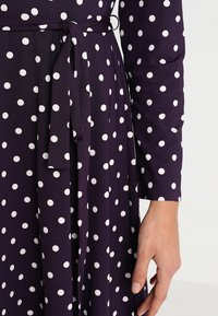 Mörklila klänning av tyg med vita prickar, knytslips i midjan, långa ärmar och en synlig hand som vilar vid sidan.