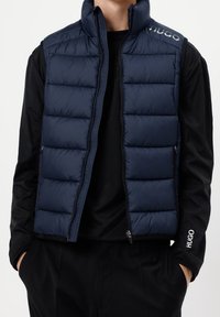 Mężczyzna w granatowej ocieplanej kamizelce na czarnej koszulce z długim rękawem z logo "HUGO" na ramieniu i rękawie, z rękami w kieszeniach.