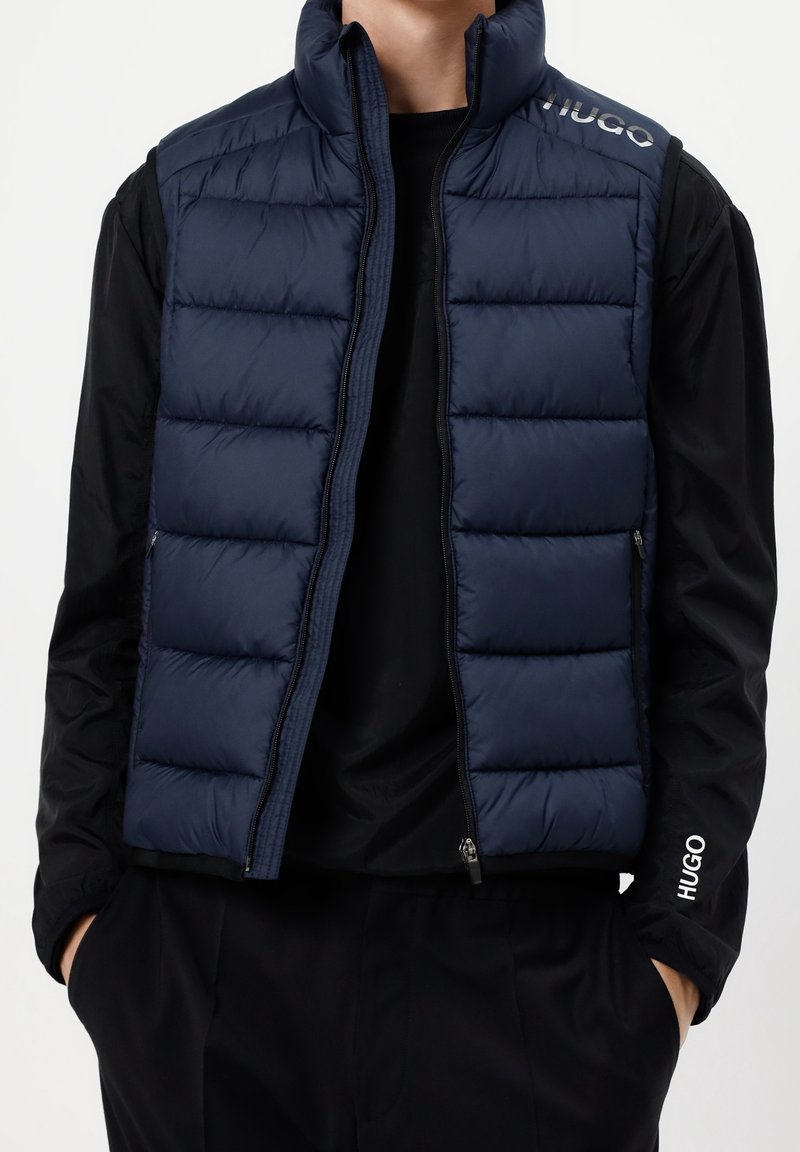 Mężczyzna w granatowej ocieplanej kamizelce na czarnej koszulce z długim rękawem z logo "HUGO" na ramieniu i rękawie, z rękami w kieszeniach.