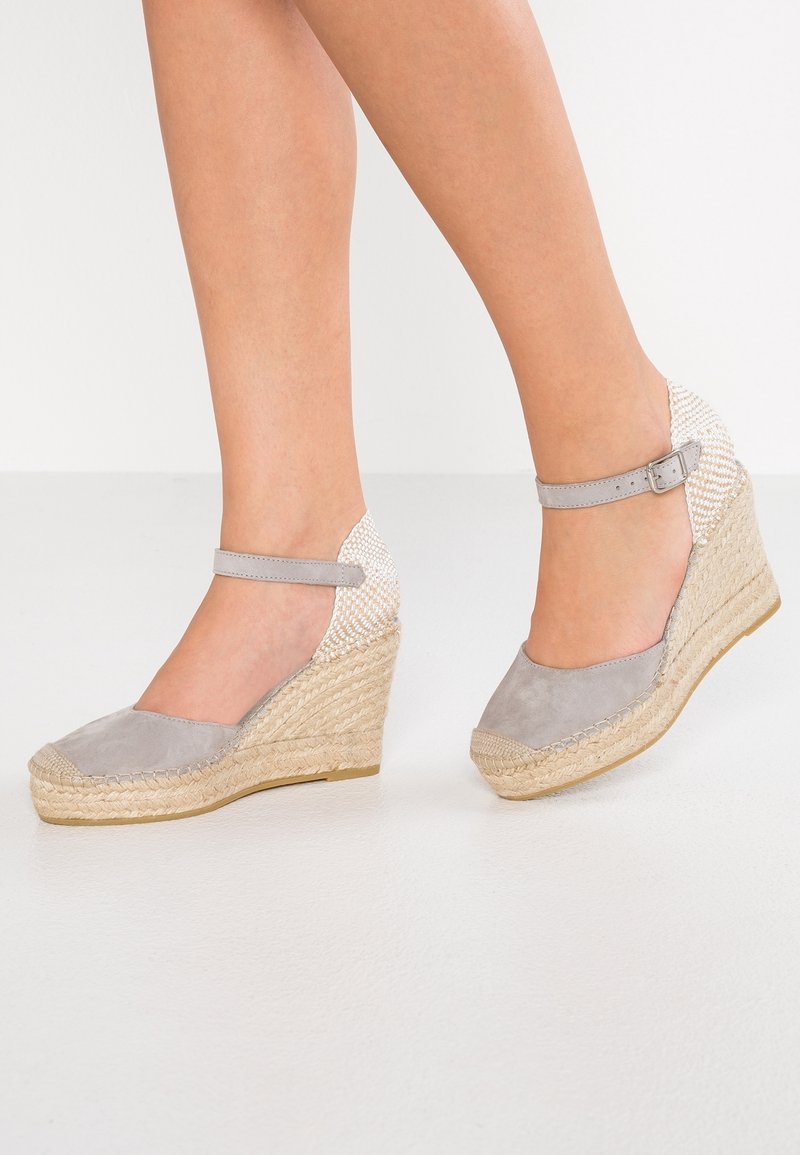 Jambes portant des espadrilles compensées gris clair avec brides à la cheville et talons en jute tissé sur un sol blanc.