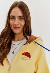 Žltá mikina na zips s hnedými akcentmi, s vyšívaným červeným croissantom a textom "petit croissant" v modrej farbe. Textúrovaná tkanina.