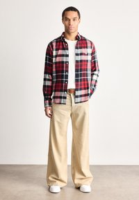 Hombre de pie con una camisa de cuadros roja, negra y blanca sobre una camiseta blanca, pantalones de pierna ancha beige, cinturón marrón y zapatillas blancas.